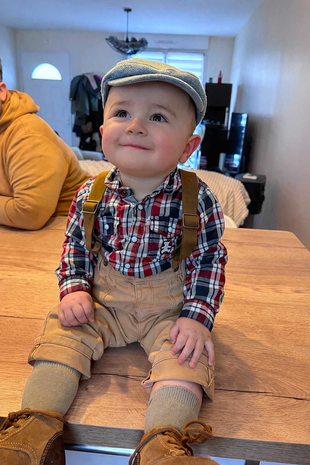 Maylan participe au concours pour gagner de l'argent avec cette photo : adult, background, child, clothing, cozy, flat_cap, furniture, happy, hoodie, indoor, person, plaid_shirt, shoes, smile, socks, suspenders, table, toddler, window, wood