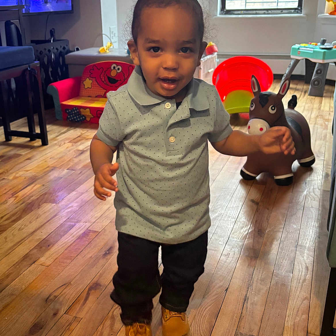 Omias is registered to the contest to win money with this photo: blinds, bouncy_horse_toy, child, curly_hair, dark_pants, elmo_chair, expression, flooring, indoor, living_room, man_on_tv, person, polo_shirt, television, toddler, toy, walking, window, wooden_floor, yellow_boots