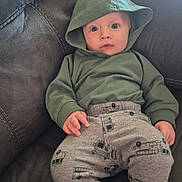 Jensen is registered to the contest to win money with this photo: baby, child, hoodie, green_clothing, gray_pants, fire_truck_pattern, couch, furniture, indoors, wide_eyes, expression, person, infant, casual_clothing, relaxing, seated, comfortable, soft_fabric, home, cute