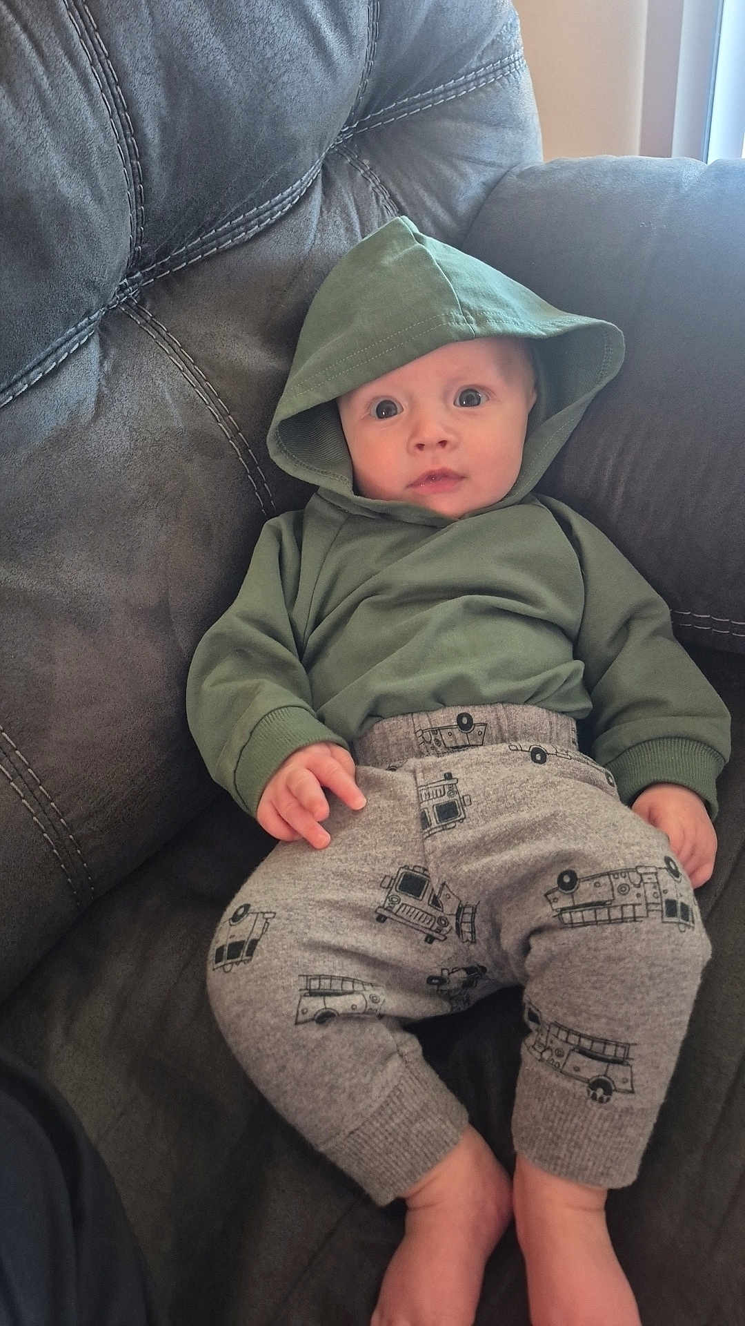 Jensen is registered to the contest to win money with this photo: baby, child, hoodie, green_clothing, gray_pants, fire_truck_pattern, couch, furniture, indoors, wide_eyes, expression, person, infant, casual_clothing, relaxing, seated, comfortable, soft_fabric, home, cute