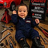 baby, child, smiling, mechanic, tools, toy_car, boots, jumpsuit, workshop, cardboard, sign, wrench, pliers, socket_wrench, floor, portrait, indoor, happy, person, infant