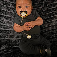 Asher joined the competition — help win amazing prizes! baby, pacifier, black_clothing, blanket, curly_hair, infant, lying_down, hand_gesture, cute, indoors, face, expression, child, soft_texture, black_boots, portrait, adorable, closeup, chain, comfortable