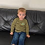 child, toddler, sitting, couch, leather, black, jeans, green_shirt, shoes, indoor, expression, portrait, young_child, furniture, casual_clothing, person, home, curious, light_hair, blue_eyes