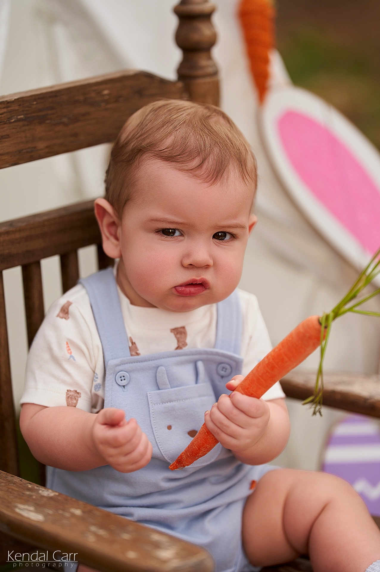 Cassidy joined the competition — help win amazing prizes! baby, child, carrot, wooden_chair, overalls, cute, serious_expression, face, hand, sitting, clothing, infant, outdoor, portrait, food, vegetable, person, plush, toddler, photography