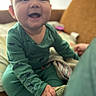 baby, child, infant, smiling, happy, sitting, portrait, face, indoors, bedroom, green_pajamas, onesie, stuffed_animal, dinosaur_toy, toy, blanket, adult, home, cozy, photograph