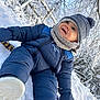 blue_clothing, child, cold_weather, daylight, face, footwear, happy, hat, mittens, nature, outdoor, playful, puffy_jacket, scarf, smiling, snow, snowy_background, toddler, trees, winter