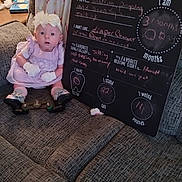 Bailey Lee joined the competition — help win amazing prizes! baby, infant, dress, gloves, headband, couch, blanket, blackboard, writing, milestones, living_room, lamp, furniture, person, sitting, cute, expression, indoor, child, young