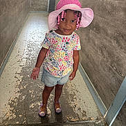 Zayla is registered to the contest to win money with this photo: child, girl, hat, pink, braids, beads, floral_shirt, denim_shorts, hallway, floor, wall, smile, shoes, indoor, portrait, cute, fashion, standing, person, colorful