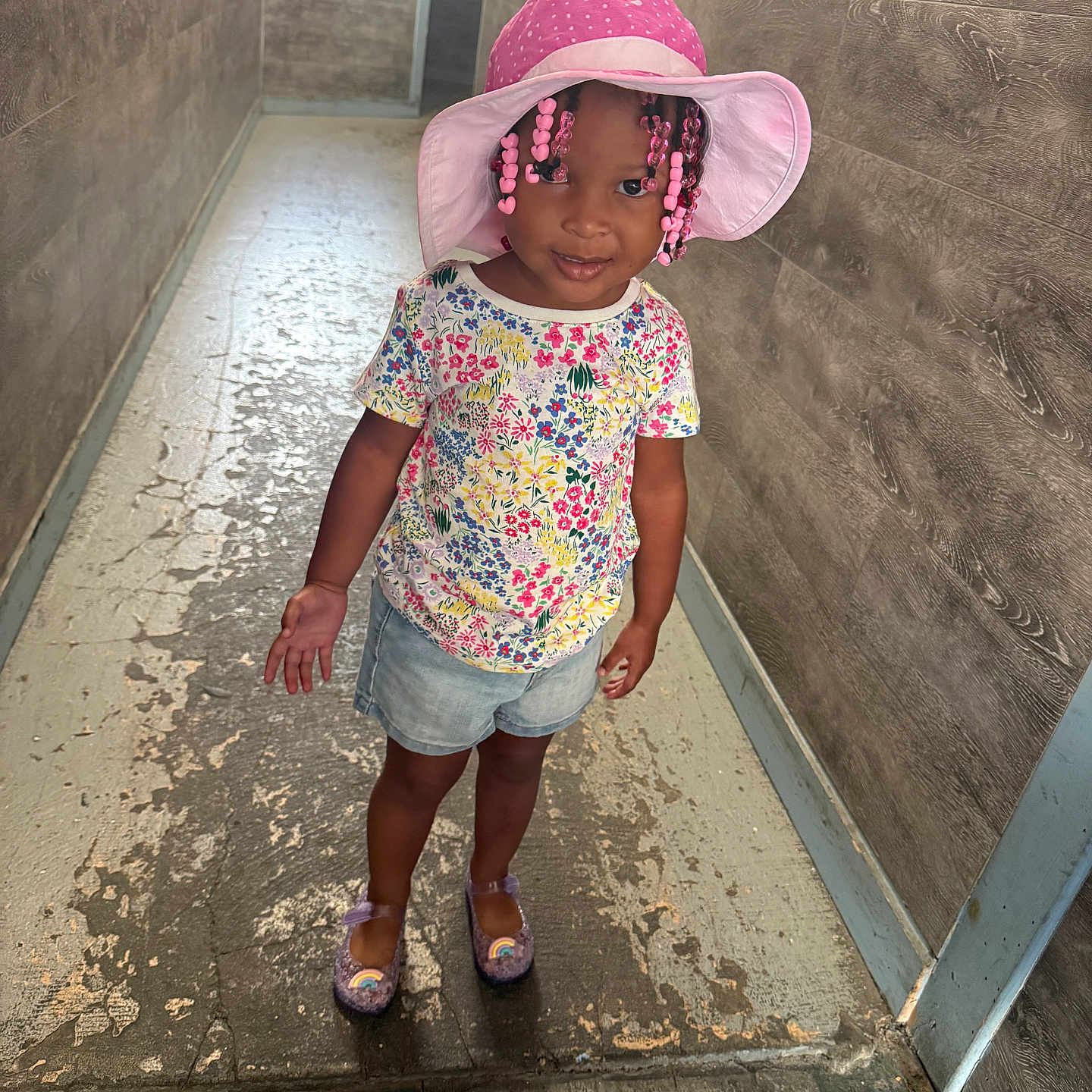 Zayla is registered to the contest to win money with this photo: child, girl, hat, pink, braids, beads, floral_shirt, denim_shorts, hallway, floor, wall, smile, shoes, indoor, portrait, cute, fashion, standing, person, colorful