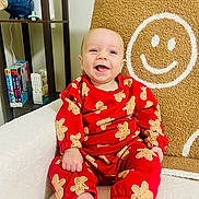 Caden is registered to the contest to win money with this photo: baby, smiling, red_clothing, gingerbread_pattern, chair, blanket, smiley_face, sitting, indoors, bookshelf, books, happy, child, cozy, cute, portrait, face, foot, hand, furniture