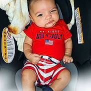 Jaxson is registered to the contest to win money with this photo: baby, infant, child, car_seat, red_clothing, striped_shorts, american_flag, patriotic, expression, serious_face, indoor, blanket, warning_label, footwear, person, sitting, portrait, cute, holiday, celebration