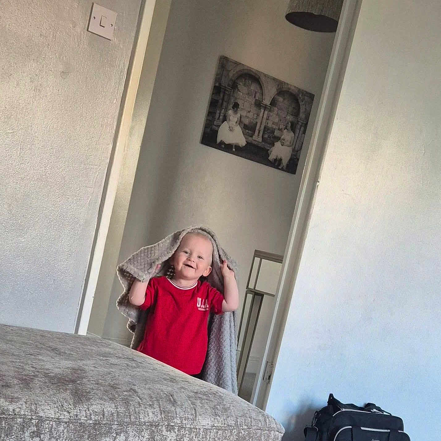 Carson joined the competition — help win amazing prizes! bag, blanket, carpet, child, cozy, cute, doorway, furniture, happy, home, indoor, light, living_room, ottoman, person, portrait, red_shirt, smiling, toddler, wall_art