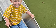 Carson is registered to the contest to win money with this photo: child, toddler, slide, playground, yellow_shirt, striped_shirt, jeans, sneakers, smiling, outdoor, metal_slide, green_ground, happy, fun, playing, casual_clothing, young_child, daylight, recreation, park