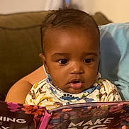 Purpose is registered to the contest to win money with this photo: baby, child, infant, reading, book, picture_book, couch, blanket, indoors, portrait, sitting, curious, focused, face, eyes, hands, colorful, cozy, family, home