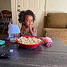 child, toddler, portrait, curly_hair, smiling, sitting, popcorn, snack, bowl, living_room, couch, table, lamp, toy, remote_control, indoors, home, door, dining_chair, movie_night