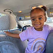 Promise is registered to the contest to win money with this photo: child, girl, toddler, smiling, portrait, car, vehicle, seated, backseat, rainbow_shirt, leopard_print, clothing, face, eyes, hair_bun, indoors, sunlight, car_seat, steering_wheel, happy