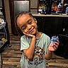 child, smiling, portrait, indoor, kitchen, snack, food_wrapper, standing, hand_on_cheek, braids, green_shirt, graphic_t_shirt, pajamas, high_chair, refrigerator, microwave, coffee_maker, wooden_floor, home, happy