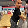 child, bowling, bowling_alley, bowling_lane, surprised, wide_eyes, open_mouth, portrait, indoor, recreation, sports, black_shirt, gray_pants, bowling_pins, bowling_ball, seating, wooden_floor, family_activity, background_people, photography