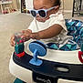 baby, sunglasses, toy, indoor, child, walker, carpet, patterned_seat, white_shirt, curly_hair, focused, hand, play, furniture, table, stool, safety, red, blue, person
