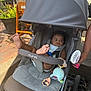 baby, stroller, child, outdoor, sunlight, foot, hand, fan, plant, pavement, person, nail_polish, toy, seat, canopy, clothing, relaxing, sidewalk, summer, shade