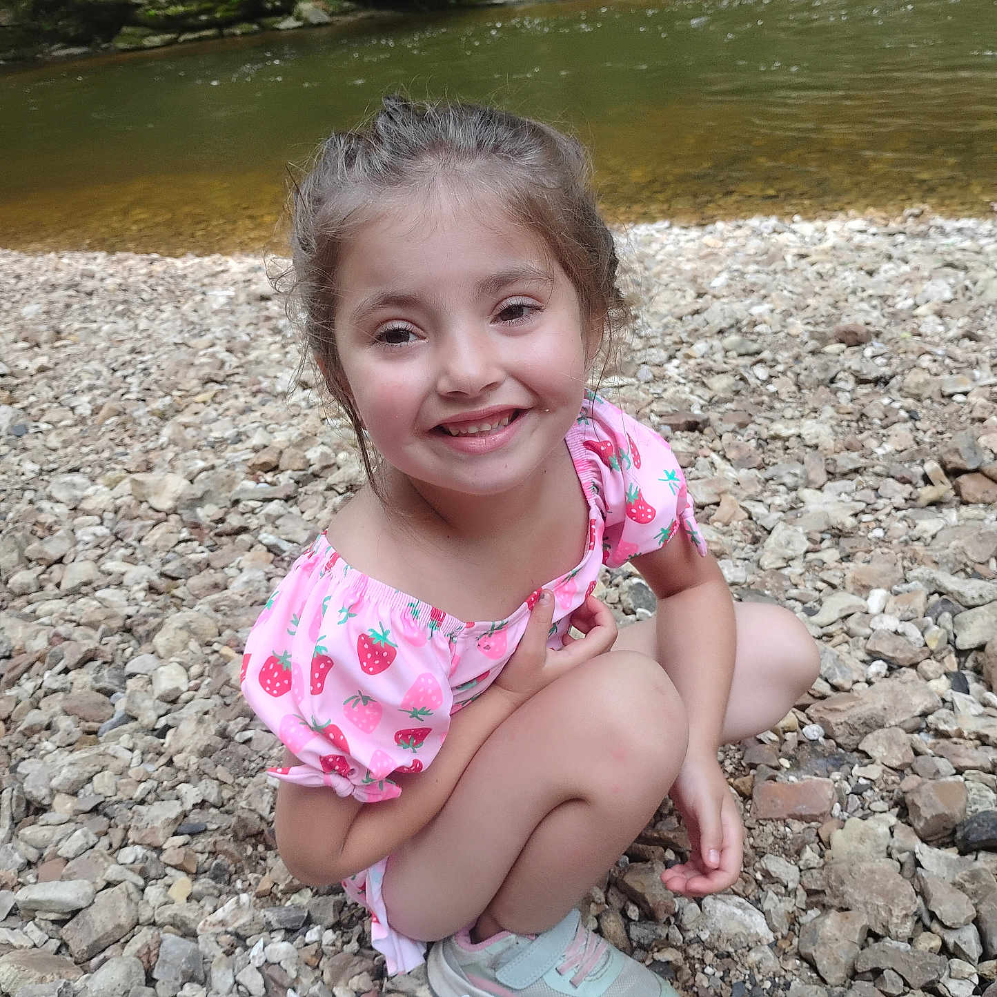 Natalie is registered to the contest to win money with this photo: child, crouching, daylight, dress, face, food, girl, hair, happy, nature, outdoor, person, picnic, pink, playful, river, rocks, smiling, snacks, sneakers