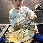 Bastien participe au concours pour gagner de l'argent avec cette photo : toddler, child, baby, face, whisk, mixing_bowl, flour, batter, kitchen, countertop, sink, stovetop, pajamas, blue_socks, electrical_outlet, electrical_plug, tile_wall, messy, baking, sitting