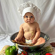 Benny is registered to the contest to win money with this photo: baby, chef_hat, spaghetti, mixing_bowl, diaper, green_bell_pepper, red_bell_pepper, tomatoes, green_onions, onion, bread, wooden_spoon, food, kitchen, cute, child, sitting, playful, white_background, healthy_food