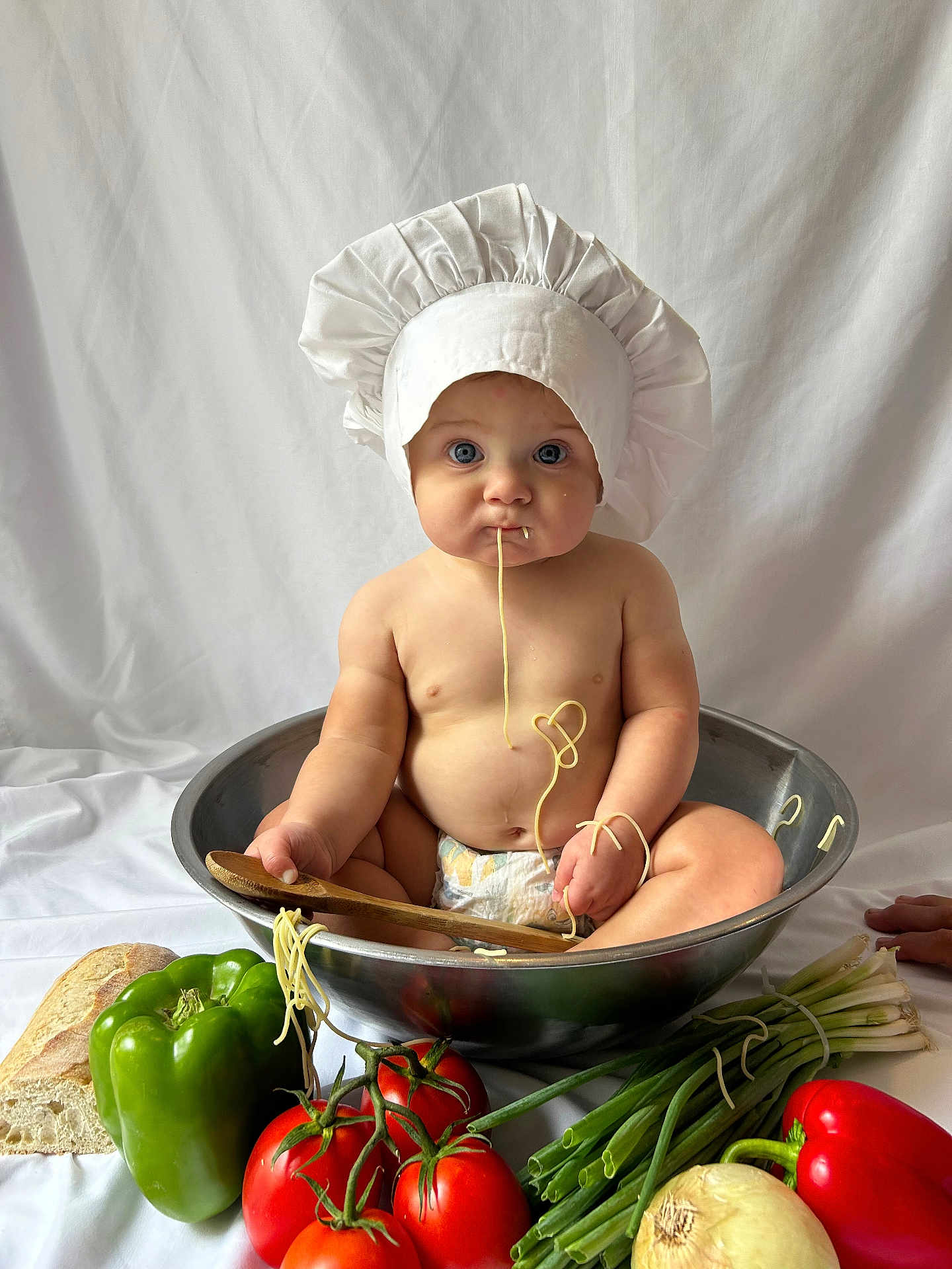 Benny is registered to the contest to win money with this photo: baby, chef_hat, spaghetti, mixing_bowl, diaper, green_bell_pepper, red_bell_pepper, tomatoes, green_onions, onion, bread, wooden_spoon, food, kitchen, cute, child, sitting, playful, white_background, healthy_food