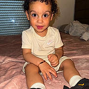 Gonçalves a rejoint le concours — aidez-le/la à gagner de superbes lots ! toddler, child, curly_hair, big_eyes, beige_clothing, nike_logo, bracelet, black_sneakers, white_socks, pink_blanket, indoor, bedroom, bed, window, curtains, smiling, sitting, young_child, portrait, person