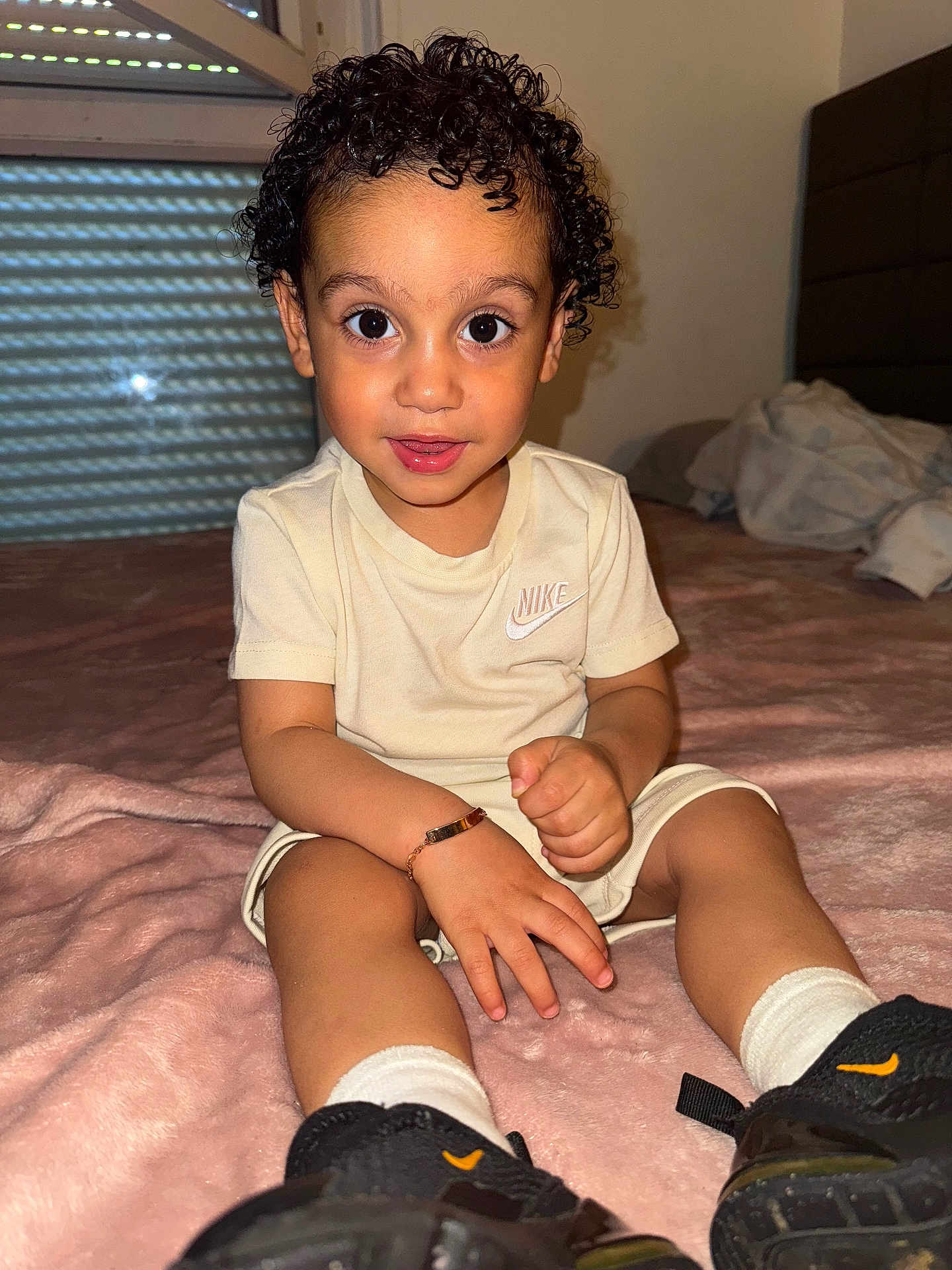 Gonçalves a rejoint le concours — aidez-le/la à gagner de superbes lots ! toddler, child, curly_hair, big_eyes, beige_clothing, nike_logo, bracelet, black_sneakers, white_socks, pink_blanket, indoor, bedroom, bed, window, curtains, smiling, sitting, young_child, portrait, person