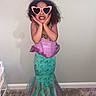 child, girl, mermaid_costume, dress, pink, green, heart_shaped_sunglasses, smiling, posing, indoor, carpet, wall, curly_hair, playful, costume, fashion, fun, cute, portrait, happy