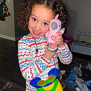 Aria is registered to the contest to win money with this photo: child, curly_hair, smiling, pajamas, stuffed_toy, pink_bird, indoor, floor, toys, wrapping_paper, holiday, happy, cute, person, face, play, colorful_clothing, home, childhood, celebration