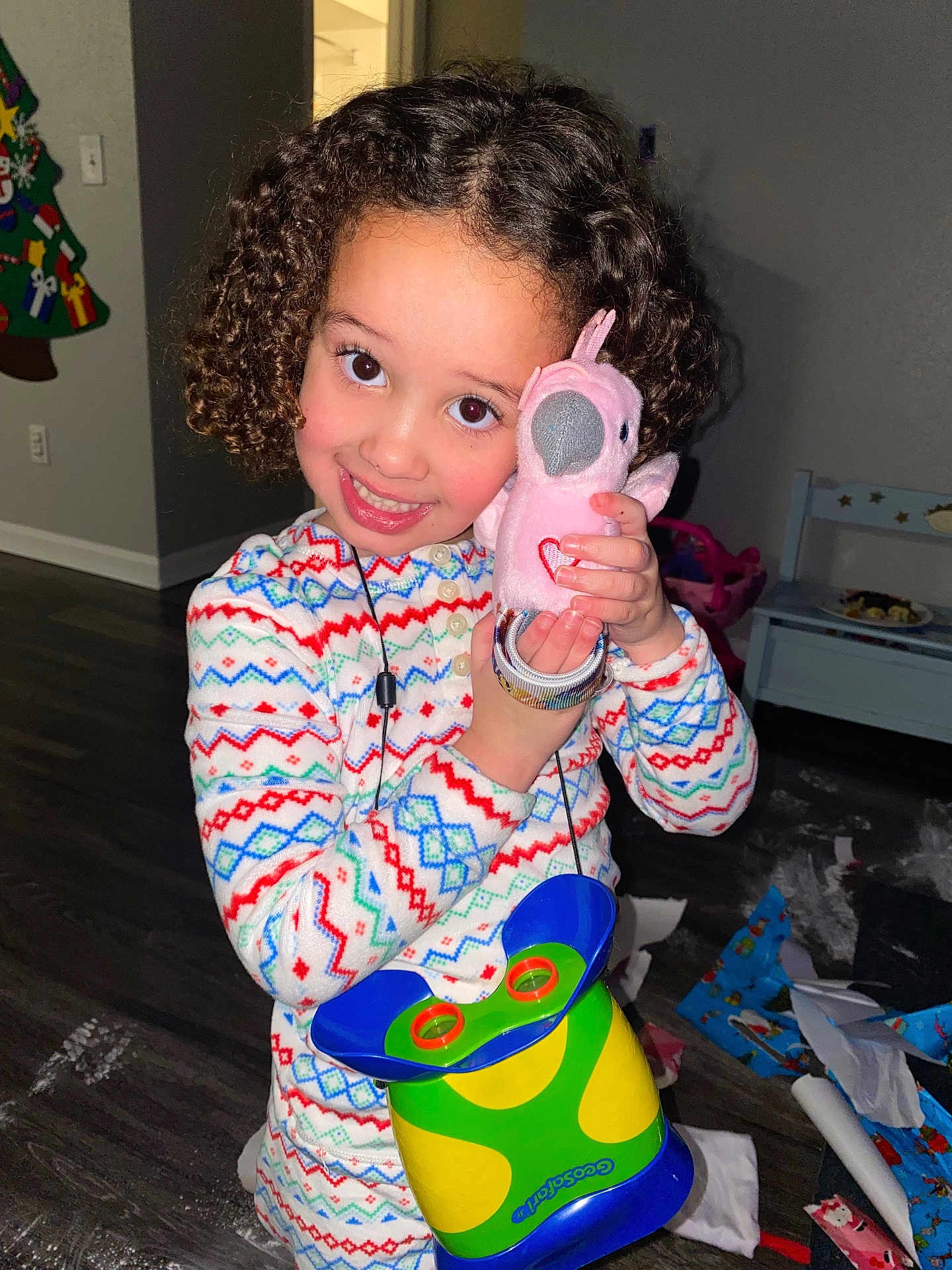 Aria is registered to the contest to win money with this photo: child, curly_hair, smiling, pajamas, stuffed_toy, pink_bird, indoor, floor, toys, wrapping_paper, holiday, happy, cute, person, face, play, colorful_clothing, home, childhood, celebration