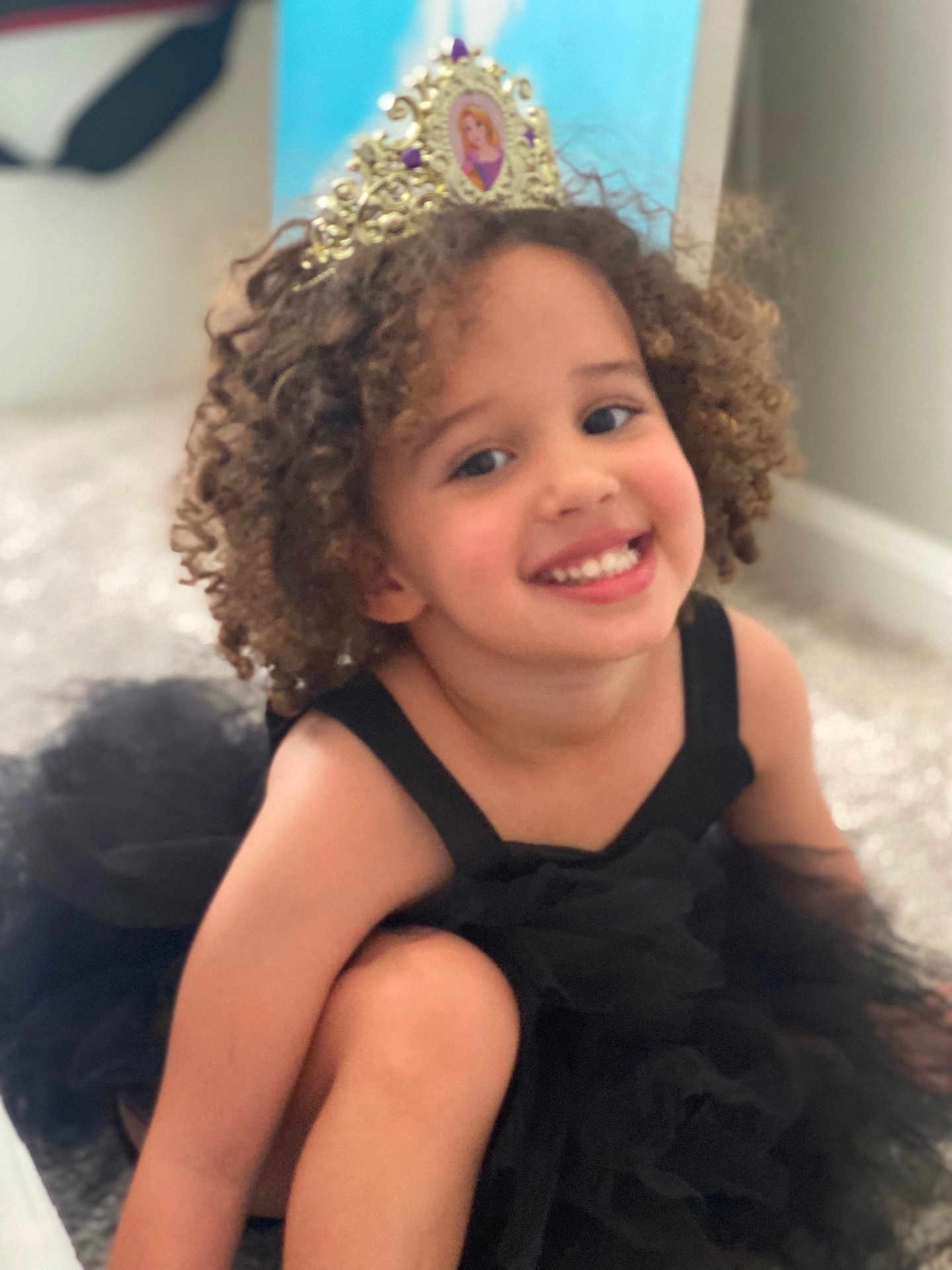 Aria is registered to the contest to win money with this photo: child, smiling, curly_hair, black_dress, tutu, tiara, princess, indoors, carpet, happy, portrait, cute, young_child, girl, playful, costume, headwear, fashion, person, sitting