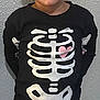 child, costume, skeleton, black_clothing, pink_heart, curly_hair, smile, standing, gray_wall, portrait, long_sleeve, pants, young, casual, indoors, cute, fashion, face, person, front_view