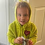 child, hoodie, green_clothing, indoor, door, curly_hair, holding_object, young_child, person, casual_wear, heart_emblem, small_box, standing, blurry, smiling, portrait, light, young_person, home