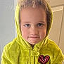 child, face, hoodie, yellow_clothing, heart_patch, indoor, portrait, curly_hair, standing, expression, person, young_child, casual_wear, wall, door, fuzzy_texture, cute, looking_at_camera, light, closeup