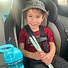 Elijah is registered to the contest to win money with this photo: child, car_seat, seatbelt, boy, cap, mama_bear, smile, holding, straws, compostable, water_bottle, blue, car_interior, window, sunlight, casual_clothing, red_shirt, gray_pants, curly_hair, happy