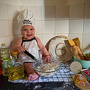 Aris joined the competition — help win amazing prizes! apron, baby, baking, bread, chef_hat, chocolate, cooking, countertop, eggs, flour, food, glass_bowl, infant, kitchen, kitchen_towel, oil_bottle, six_months, smiling, whisk, wooden_spoons