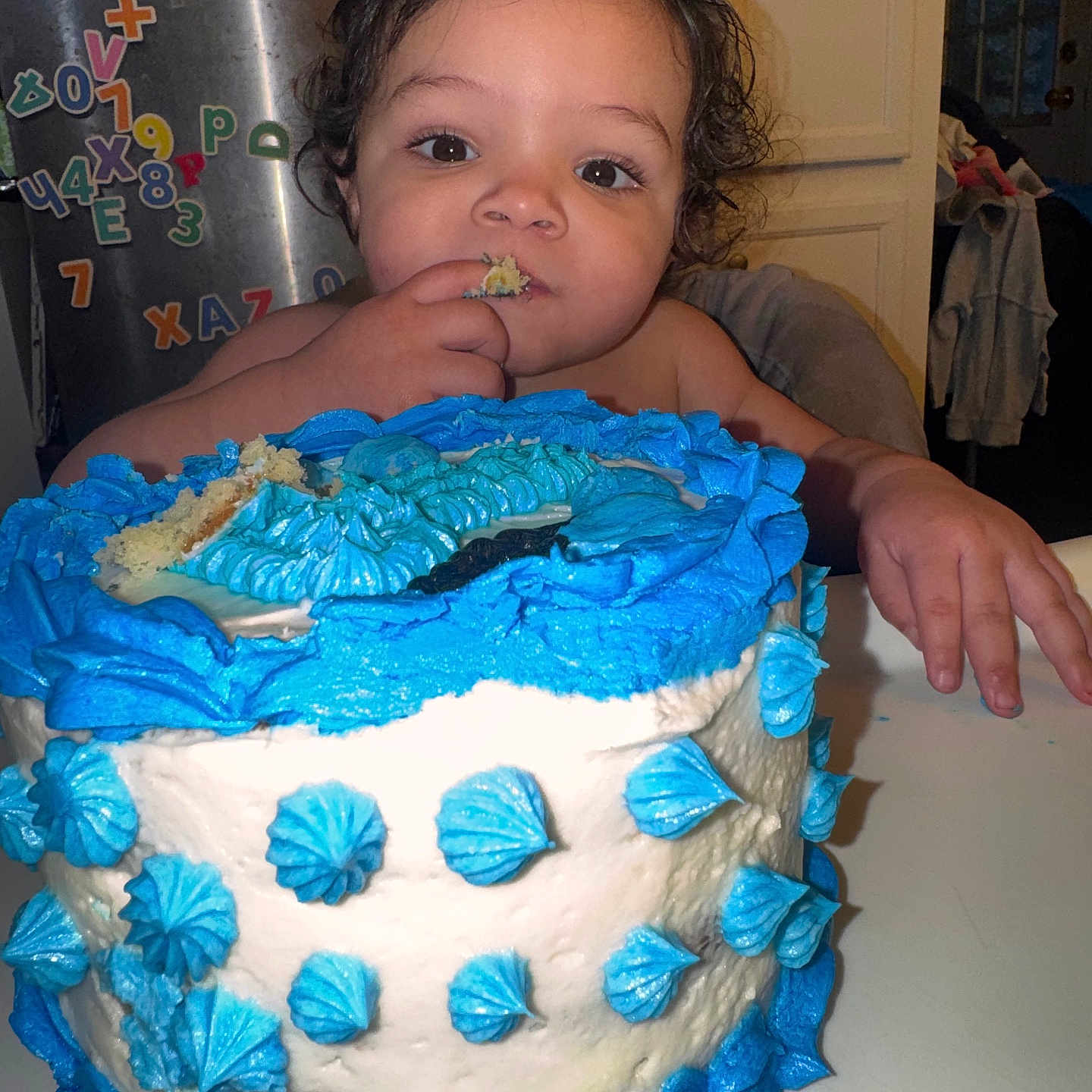 Xavier is registered to the contest to win money with this photo: child, toddler, baby, birthday, cake, frosting, blue_frosting, eating, smiling, face, portrait, indoor, kitchen, refrigerator, alphabet_magnets, table, dessert, celebration, home, photograph