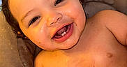 Xavier is registered to the contest to win money with this photo: baby, child, infant, smiling, happy, laughing, portrait, face, eyes, teeth, curly_hair, sitting, cushion, chair, indoor, warm_lighting, bare_chest, food, plate, closeup