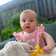 Lula is registered to the contest to win money with this photo: baby, child, pink_clothing, toy, yellow_toy, outdoor, garden, fence, curious, wide_eyes, infant, sitting, hand, portrait, ruffled_sleeves, soft_toy, blurred_background, cute, person, summer