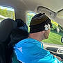 man, dog, car_interior, beanie, hoodie, driving, seat, window, daylight, black_dog, glasses, chain_necklace, headrest, greenery_outside, casual_clothing, side_view, pet, companion, relaxed, road_trip