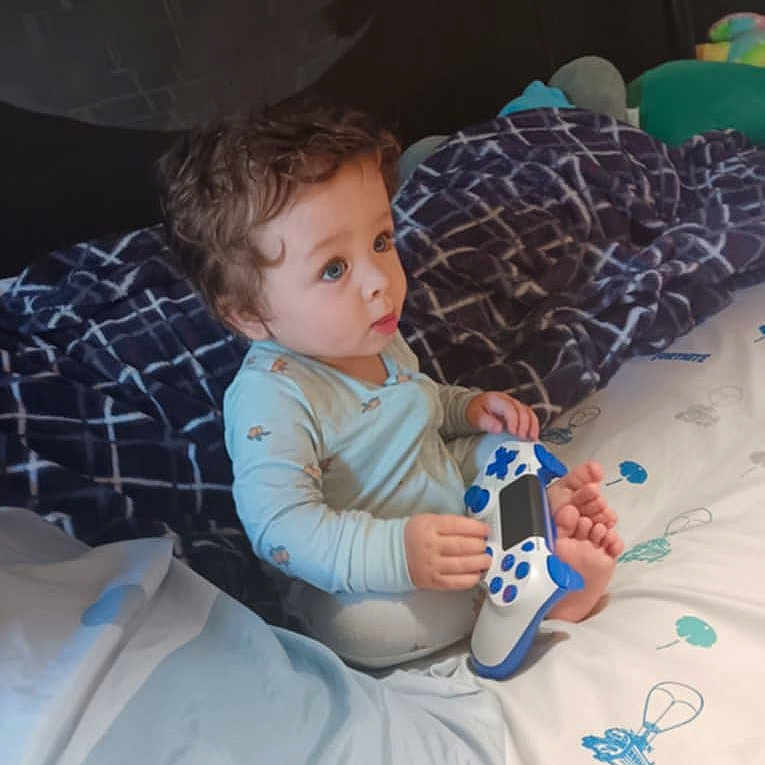 Tevy is registered to the contest to win money with this photo: bed, blanket, blue, casual, child, clothing, curly_hair, cute, foot, gaming_controller, indoors, pattern, person, play, room, sitting, toddler, toy, white, young_child