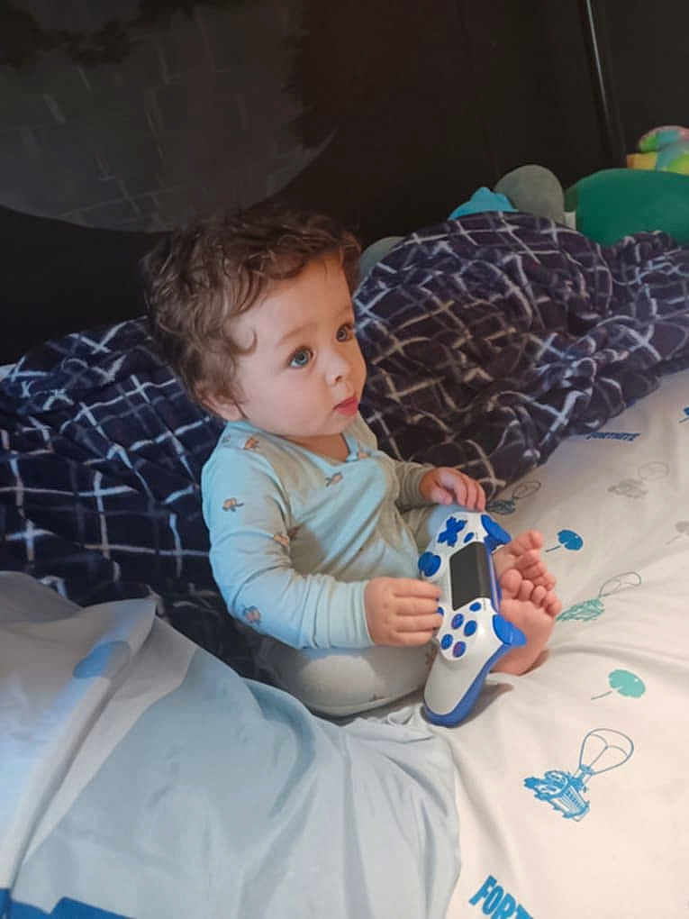 Tevy is registered to the contest to win money with this photo: toddler, child, curly_hair, bed, blanket, gaming_controller, blue, white, foot, sitting, indoors, clothing, pattern, toy, young_child, person, cute, play, room, casual