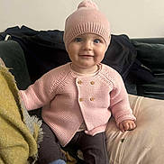 Victoire a rejoint le concours — aidez-le/la à gagner de superbes lots ! baby, child, pink_hat, pink_sweater, blue_socks, brown_pants, smile, sitting, couch, cushion, blanket, indoor, face, happy, knitted_clothing, cozy, portrait, cute, infant, warm