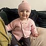 baby, child, pink_hat, pink_sweater, blue_socks, brown_pants, smile, sitting, couch, cushion, blanket, indoor, face, happy, knitted_clothing, cozy, portrait, cute, infant, warm