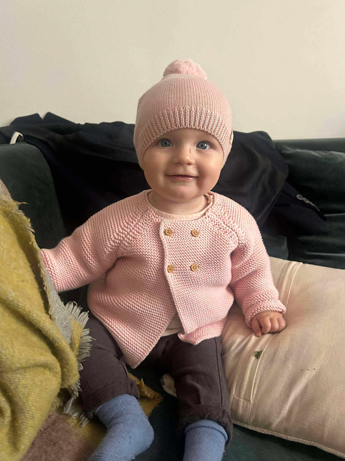 Victoire a rejoint le concours — aidez-le/la à gagner de superbes lots ! baby, child, pink_hat, pink_sweater, blue_socks, brown_pants, smile, sitting, couch, cushion, blanket, indoor, face, happy, knitted_clothing, cozy, portrait, cute, infant, warm