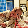 baby, infant, smiling, face, hand, blanket, striped, blue_tiles, indoor, clothing, person, cute, young_child, lying_down, soft_toys, warm_lighting, head, eyes, cheeks, pajamas