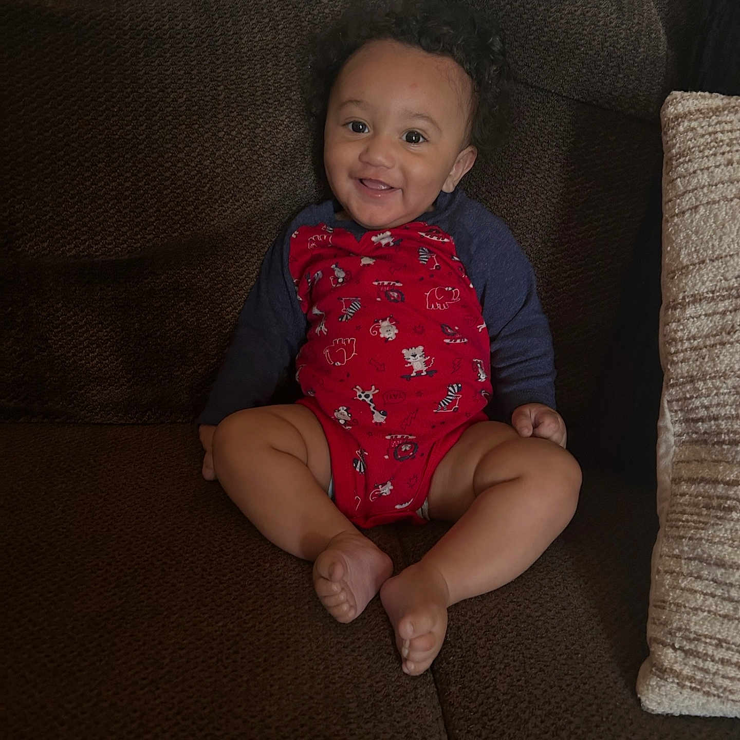 Kenzy is registered to the contest to win money with this photo: animal_prints, baby, barefoot, brown_couch, child, comfortable, couch, cozy, curly_hair, cute, happy, home, indoor, infant, plush, portrait, red_onesie, sitting, smiling, toddler